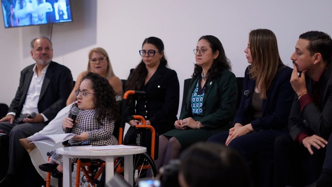 A woman in a wheelchair speaks into a microphone during a panel discussion, looking at notes on a small white table. Behind her, six other participants in professional attire sit in a row, listening attentively. The setting is a brightly lit indoor event with a plain white background.